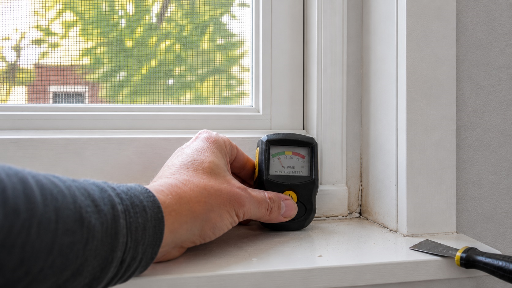 Hand holding a moisture meter against a stained lower window frame with cracked caulk and a putty knife nearby.