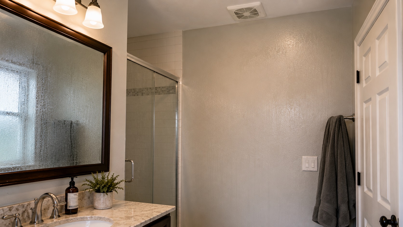 A clean bathroom after shower use with a fogged mirror, subtle condensation on the painted wall, a closed door, towel, and ceiling exhaust fan showing lingering humidity.