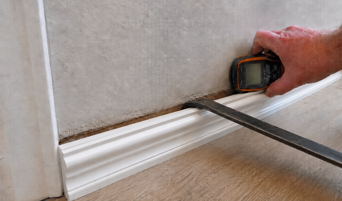 A homeowner uses a moisture meter while prying a white baseboard away from the wall to check for hidden moisture near the floor.