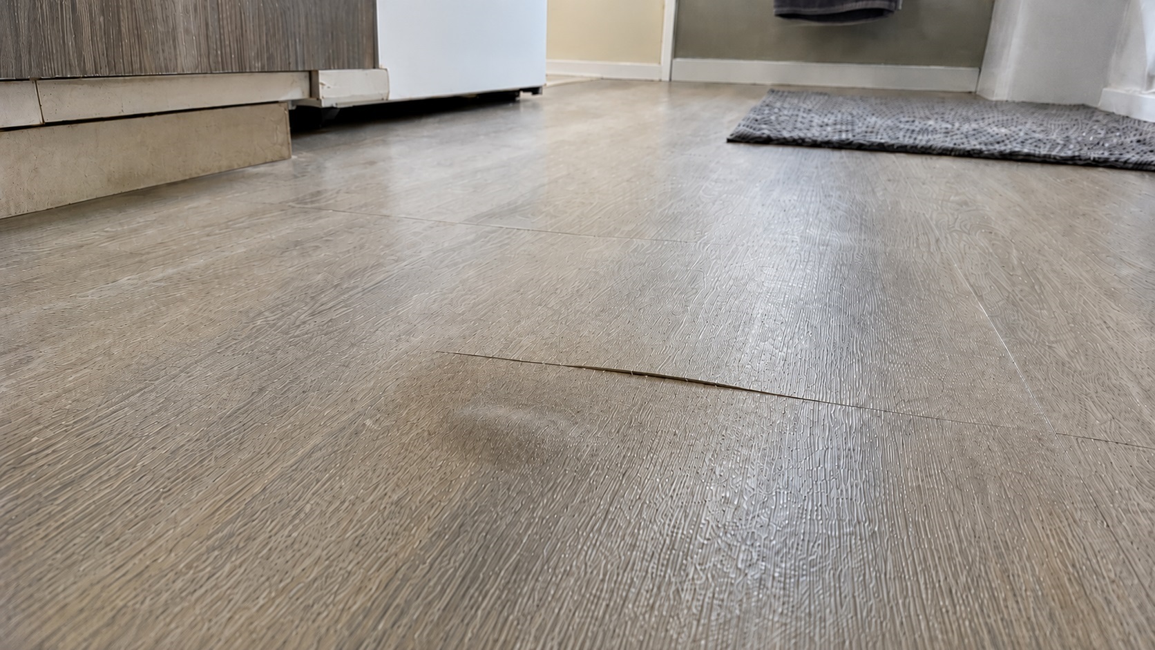 Low-angle view of vinyl plank flooring with a slightly lifted seam, dull patch, and subtle discoloration suggesting moisture trapped underneath.