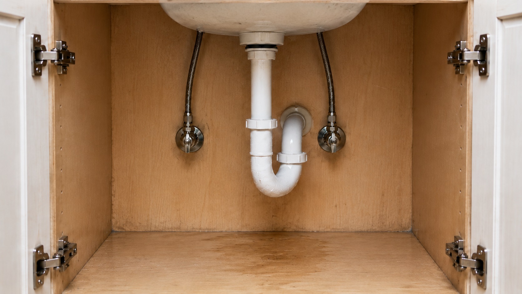 Early water damage stain under a bathroom sink cabinet with a white PVC P-trap, supply lines, and light cabinet wear.