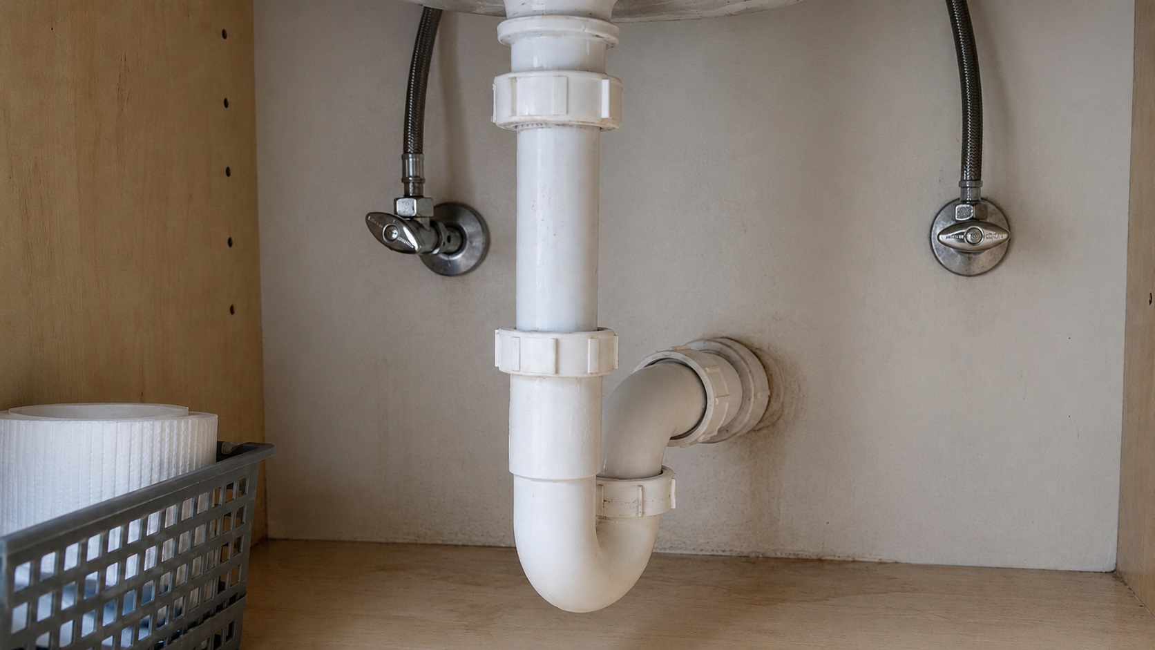 Realistic under-sink kitchen cabinet plumbing with a P-trap, shutoff valves, braided supply lines, and subtle moisture staining near hidden leak-prone connections.