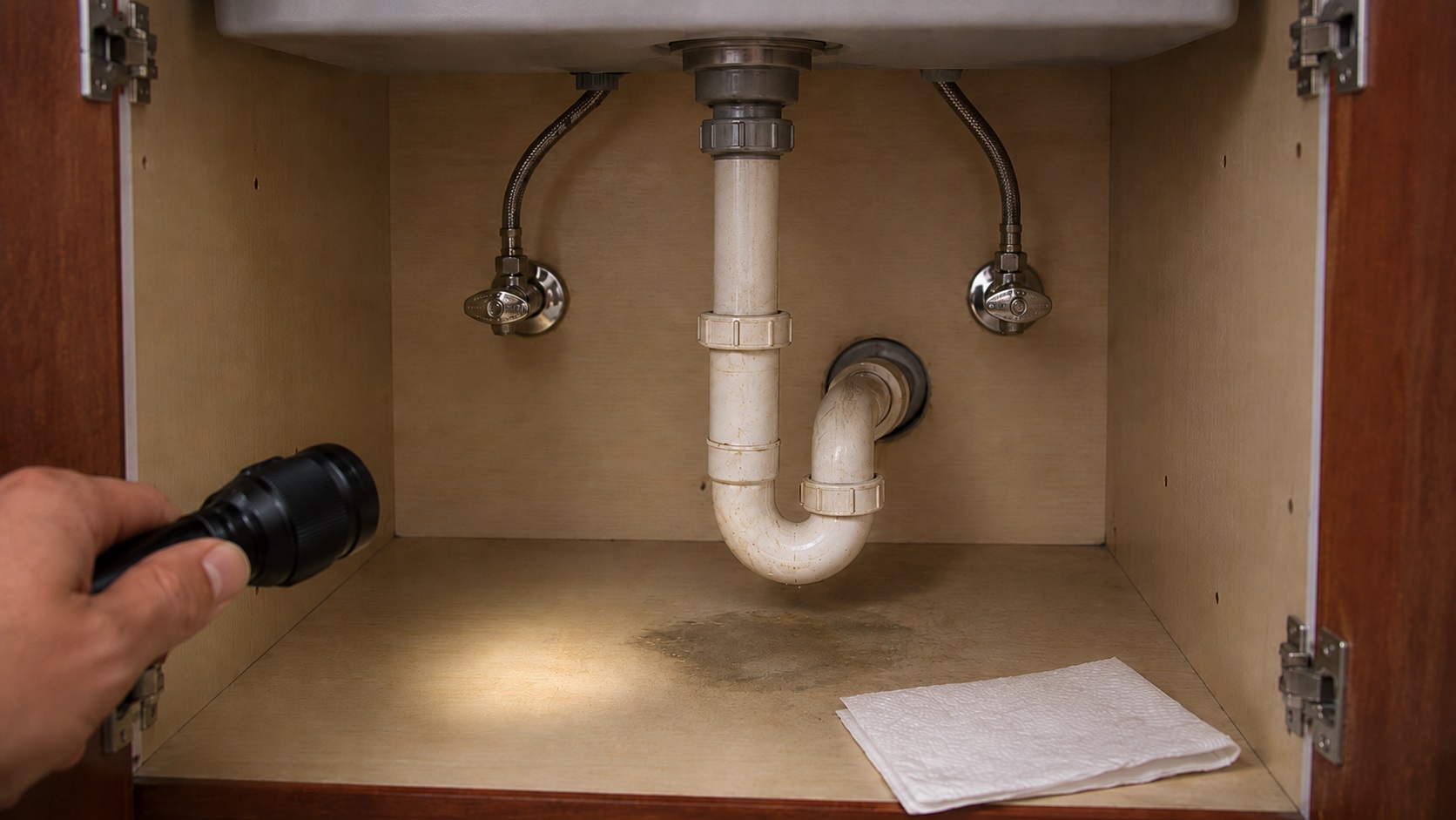 Open under-sink cabinet with realistic drain plumbing, supply lines, flashlight inspection, paper towel, and faint moisture staining on the cabinet floor.