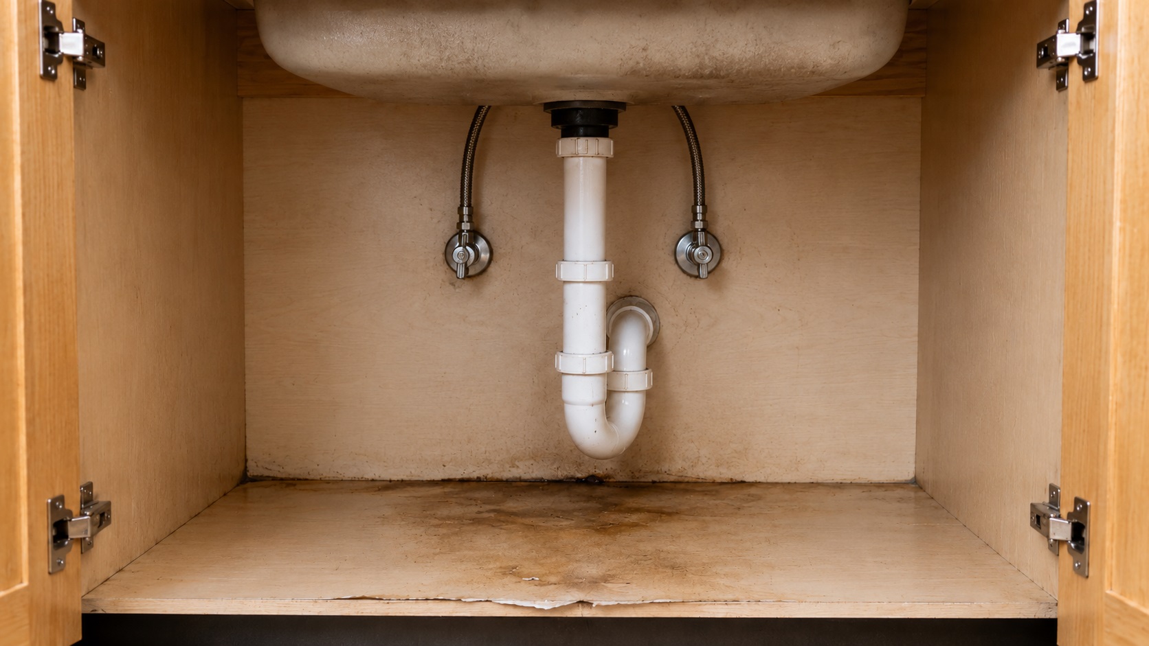 Open kitchen sink cabinet with centered plumbing, supply lines, stained cabinet floor, and darkened seams showing signs of water damage under the sink.