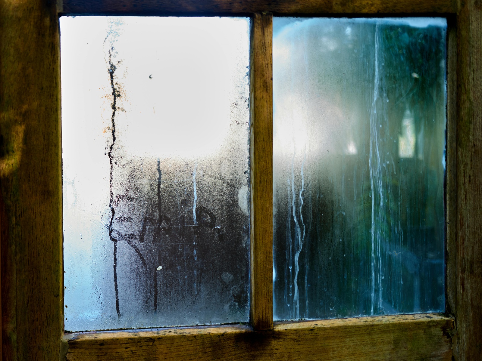 Condensation and water streaks on an old wooden window caused by moisture collecting on cold glass.