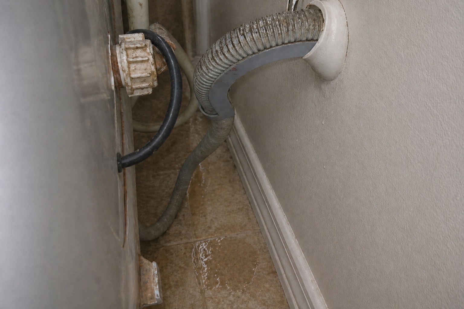 Small hidden water puddle behind washing machine near supply hoses in a realistic laundry room leak scenario