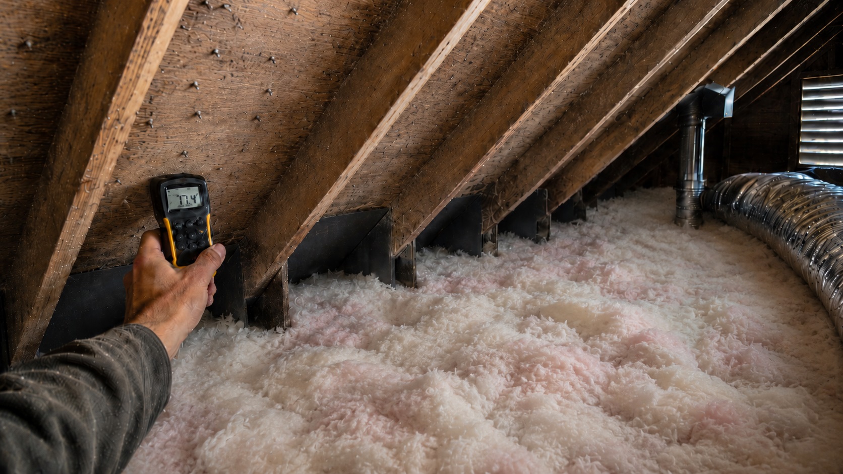 Contractor using a moisture meter on stained attic roof sheathing near ventilation baffles, ductwork, exposed rafters, roofing nails, and blown-in fiberglass insulation.