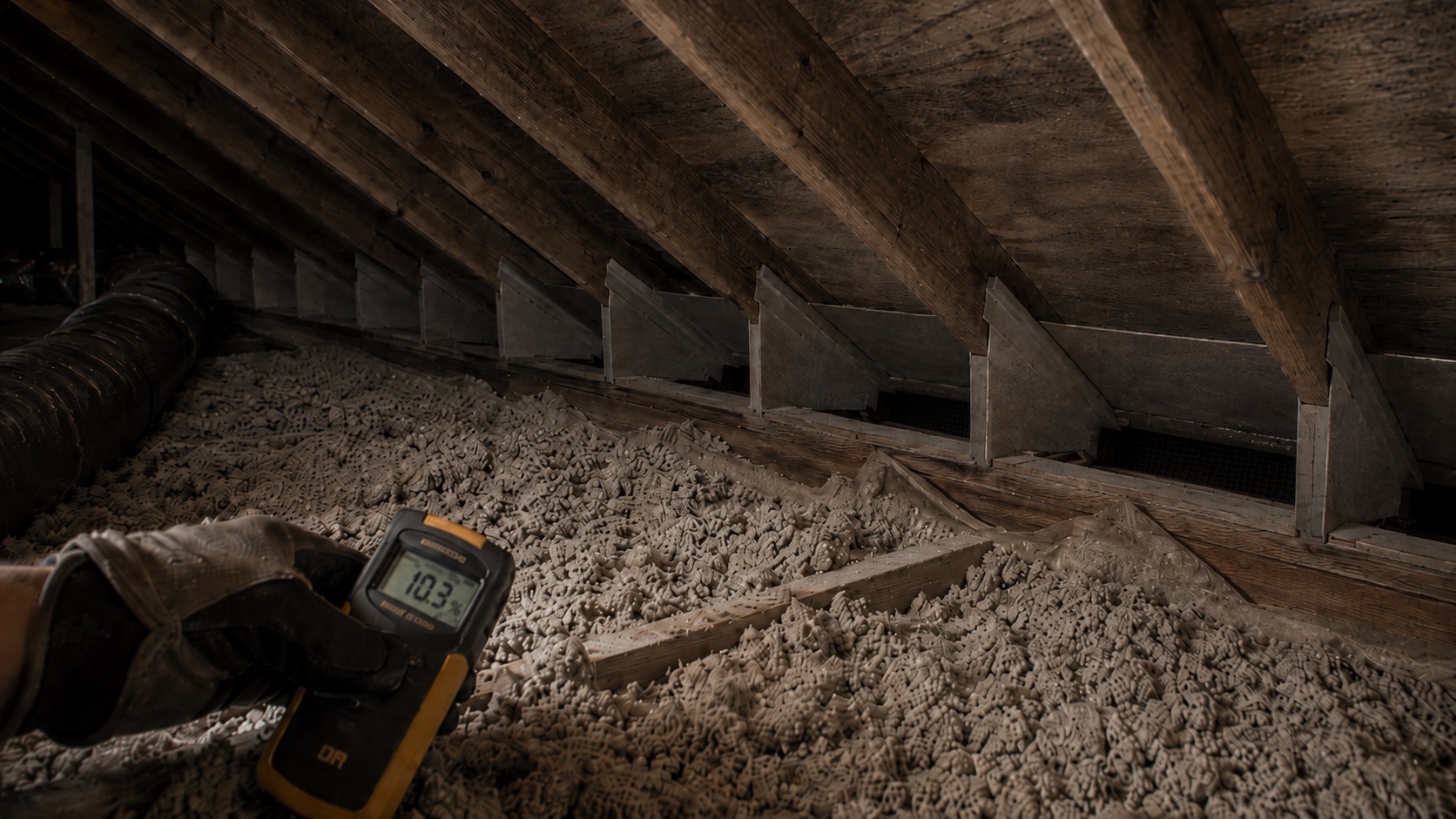 Dusty attic with eave baffles, pulled-back insulation, dry stained roof sheathing, and a moisture meter checking repairs after recurring condensation.