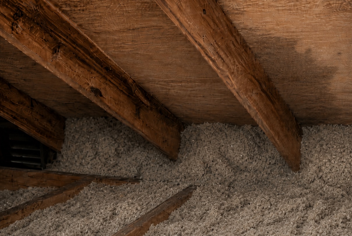 Attic insulation packed against the eave area beneath roof rafters, with faint condensation staining on the roof sheathing from poor ventilation.