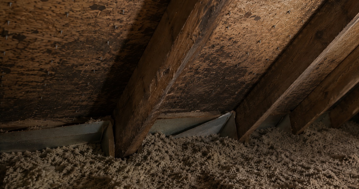 Older attic roof sheathing with roofing nail tips, faint moisture staining, and loose-fill insulation blocking a vent baffle near the eaves.