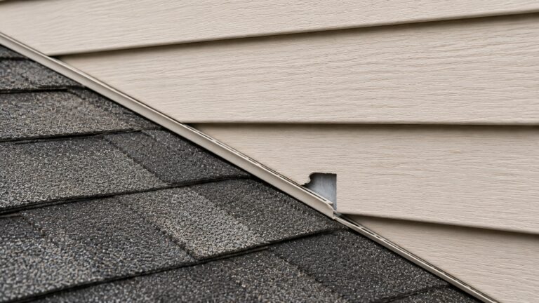 Cracked siding and J-channel above a roof edge exposing flashing at a roof-to-wall junction