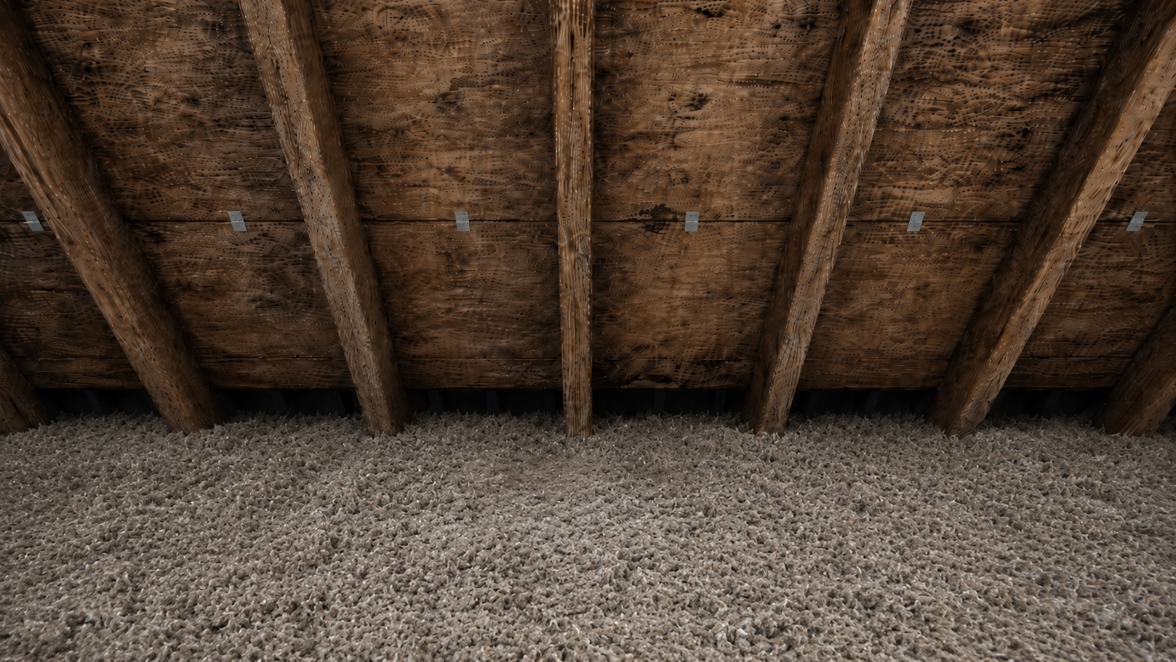 Aged attic roof decking with rafters, visible H-clips, and blown-in insulation with a subtle damp patch below the roof seam.