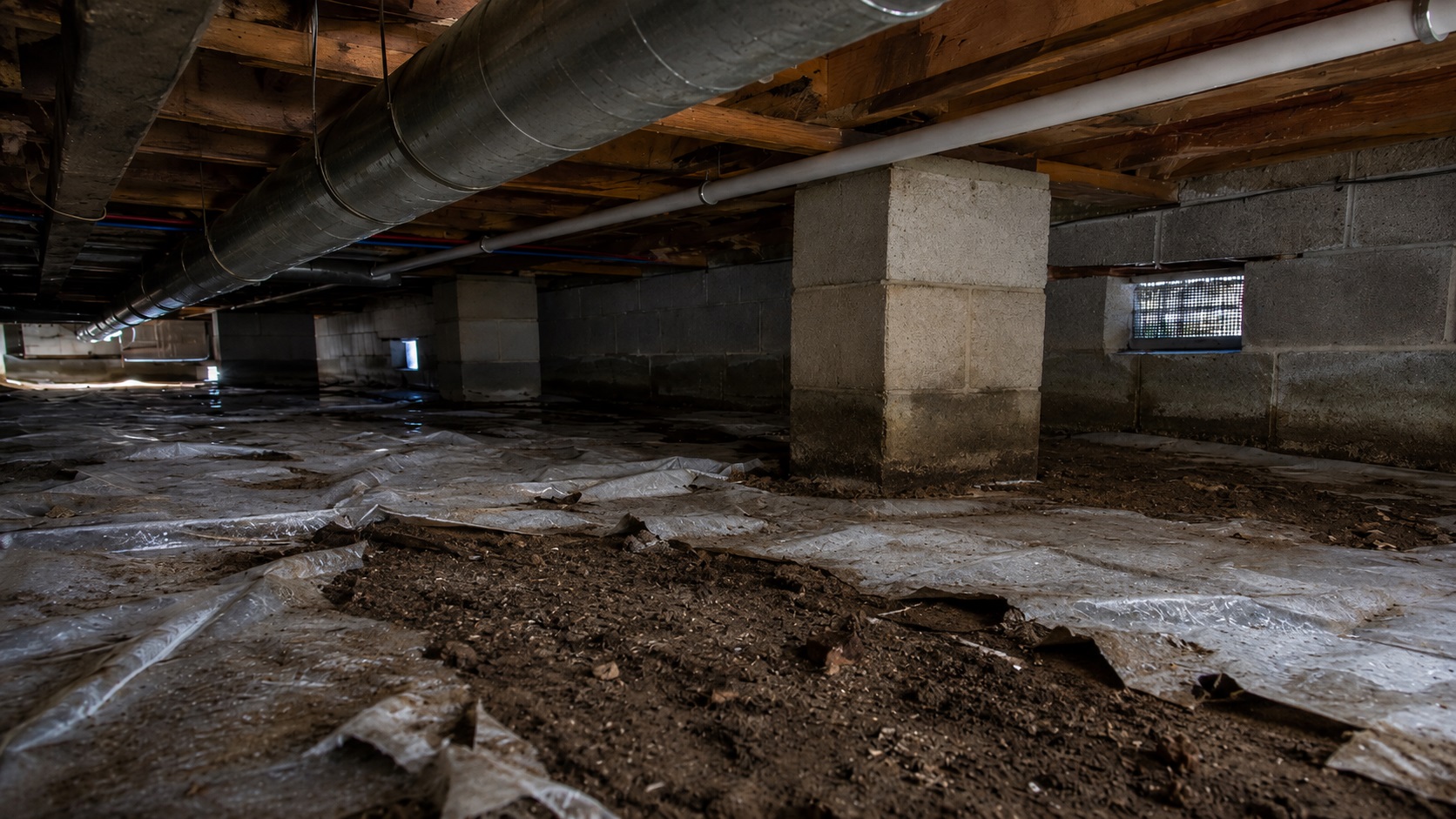 Low view of a damp crawl space with torn vapor barrier, exposed soil, concrete block piers, foundation vents, pipes, and mild moisture staining.