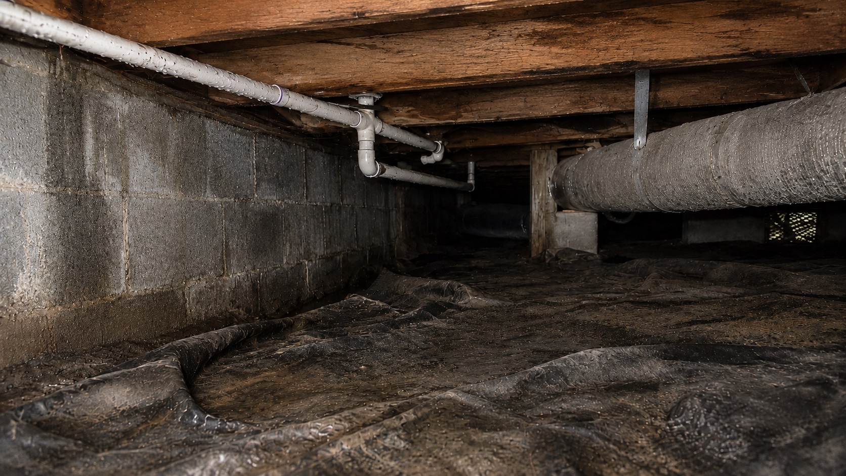 Damp crawl space with a wrinkled vapor barrier, stained block wall, exposed joists, plumbing pipes, ductwork, and subtle condensation.