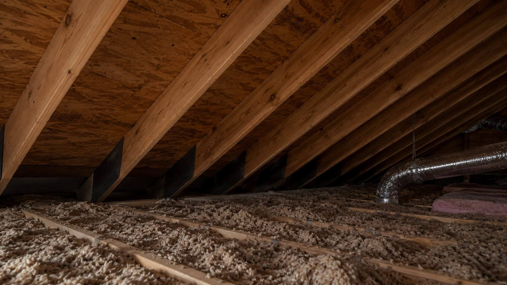 Dry attic roof framing with ventilation baffles, settled insulation, roof sheathing, and a shadowed eave airflow path.