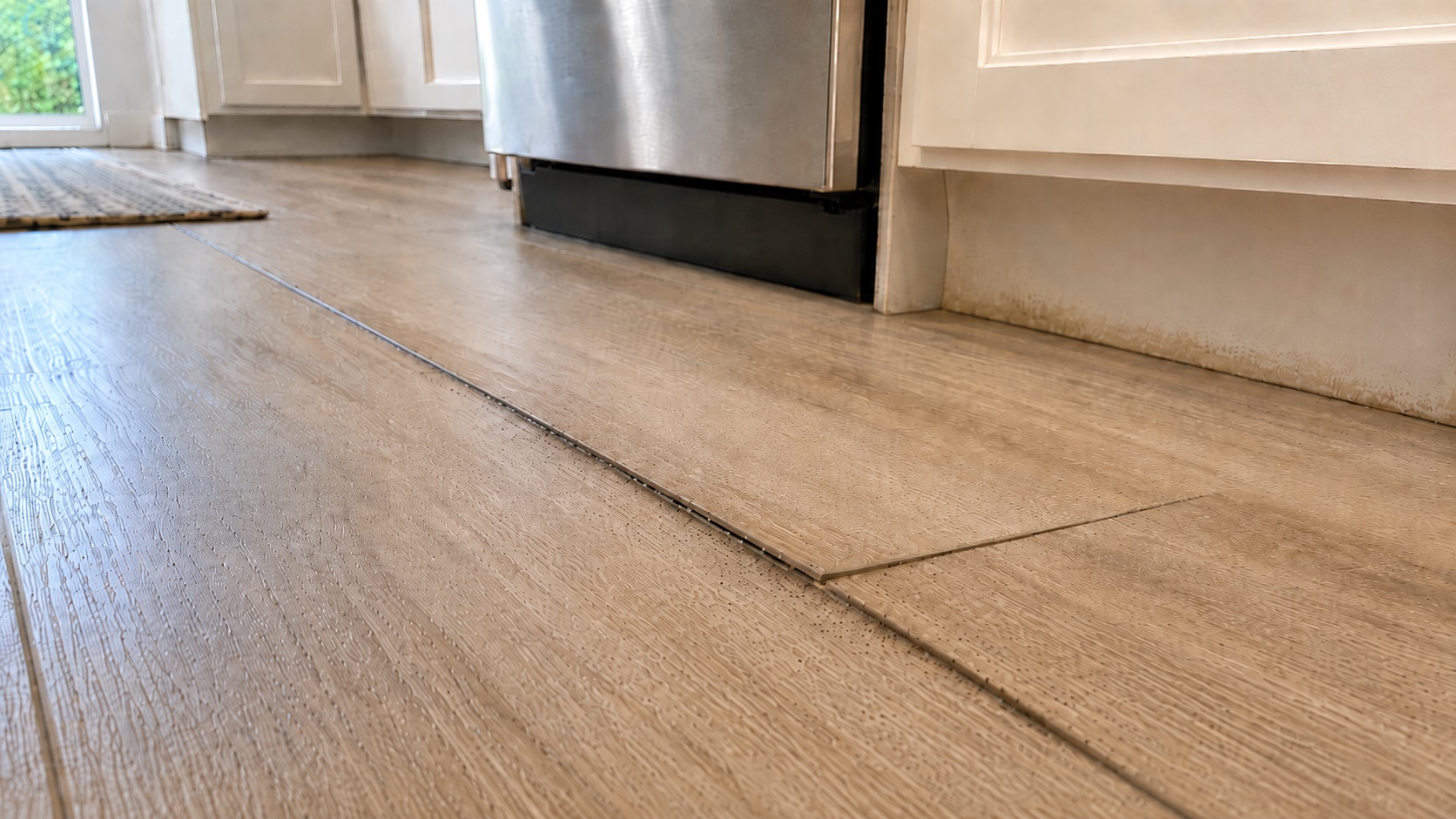 Low-angle view of a kitchen floor showing a slightly lifted plank seam with faint dark specks and subtle moisture staining near a cabinet toe-kick.