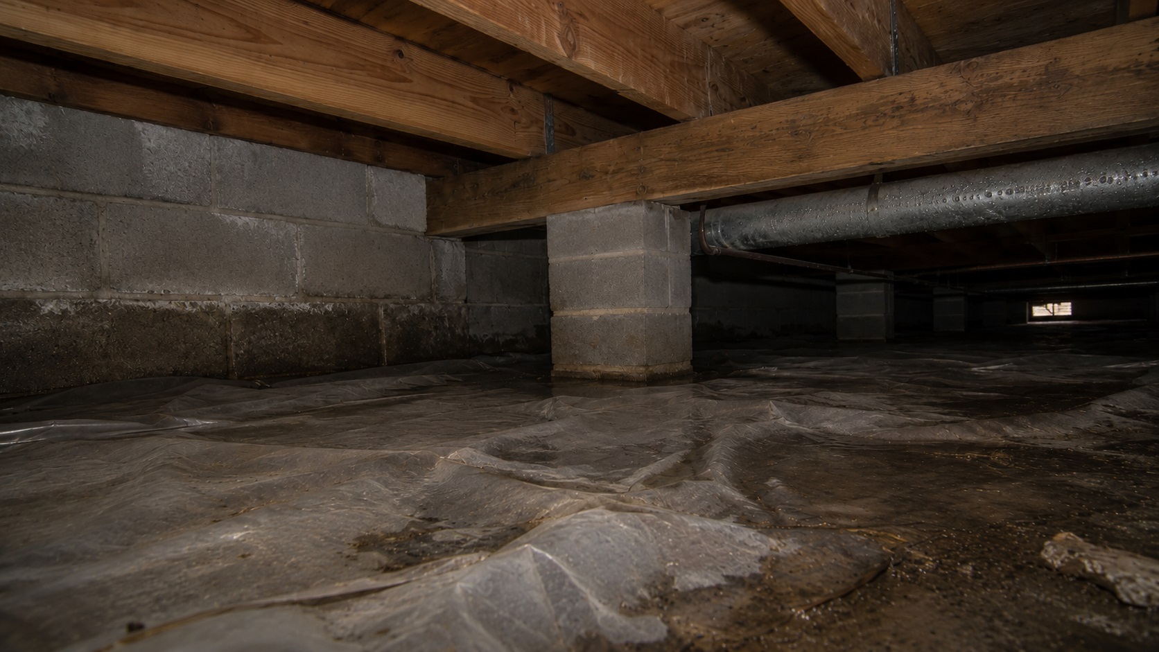 Low crawl space with wrinkled vapor barrier, damp patches, stained concrete block wall, support piers, wood joists, and condensation on a metal duct.