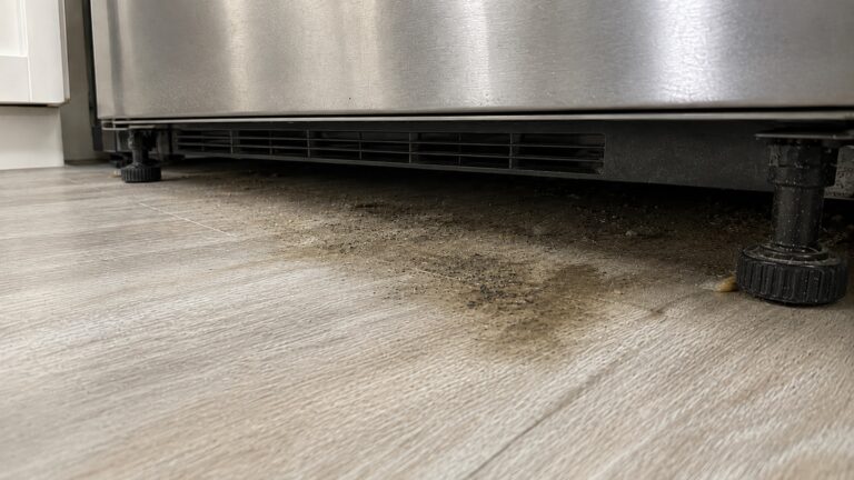 Low-angle view beneath a stainless steel refrigerator showing dust, crumbs, faint dark speckling, and a dull damp patch on the floor.