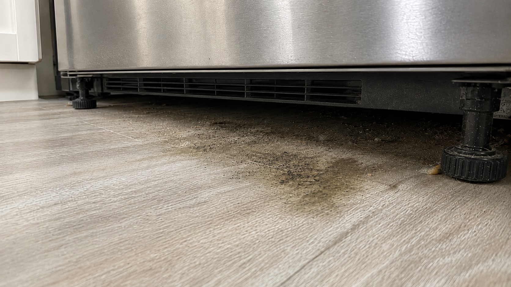 Low-angle view beneath a stainless steel refrigerator showing dust, crumbs, faint dark speckling, and a dull damp patch on the floor.