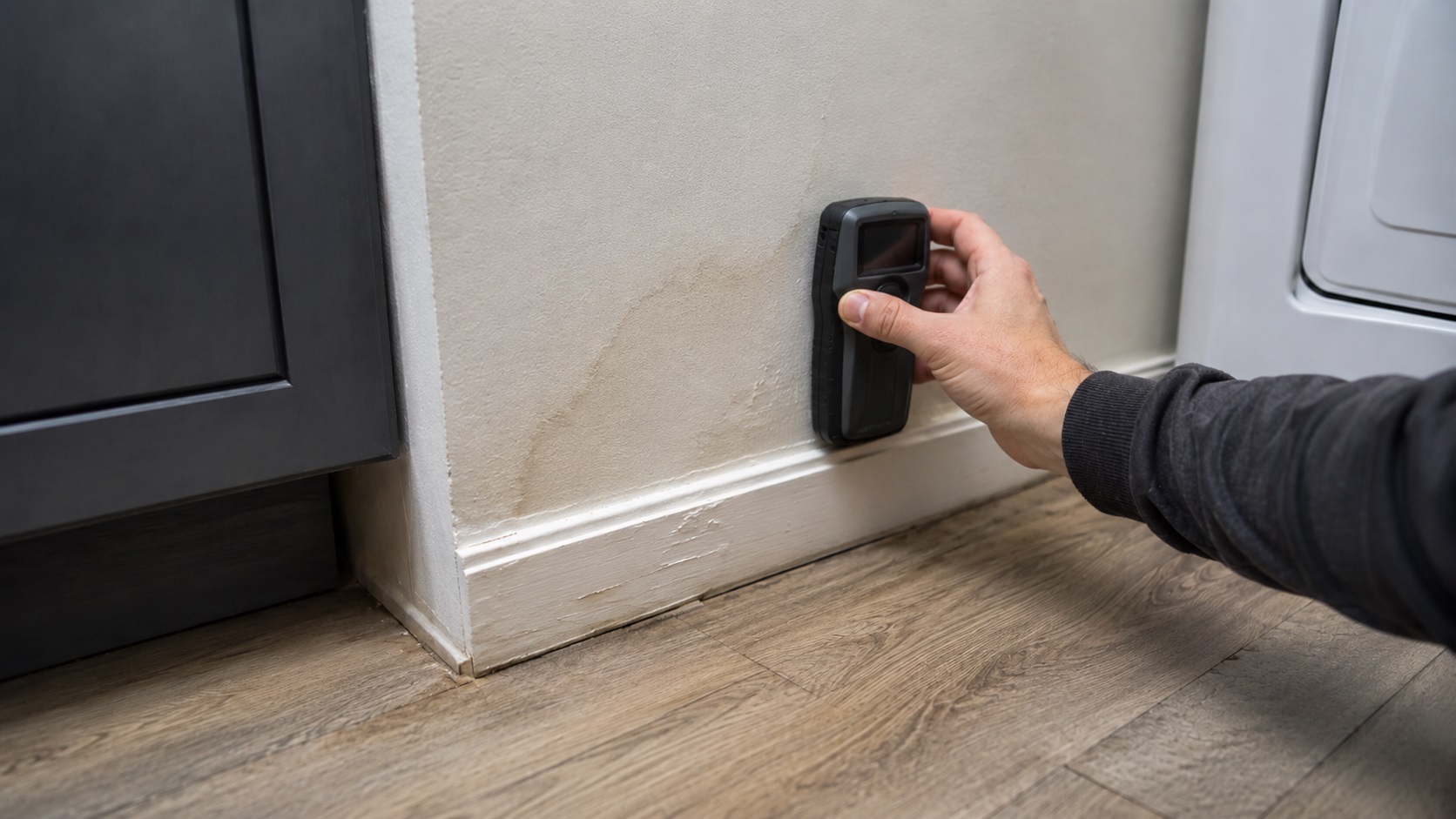 A homeowner presses a pinless moisture meter against a stained lower wall above a moisture-damaged baseboard and flooring edge.