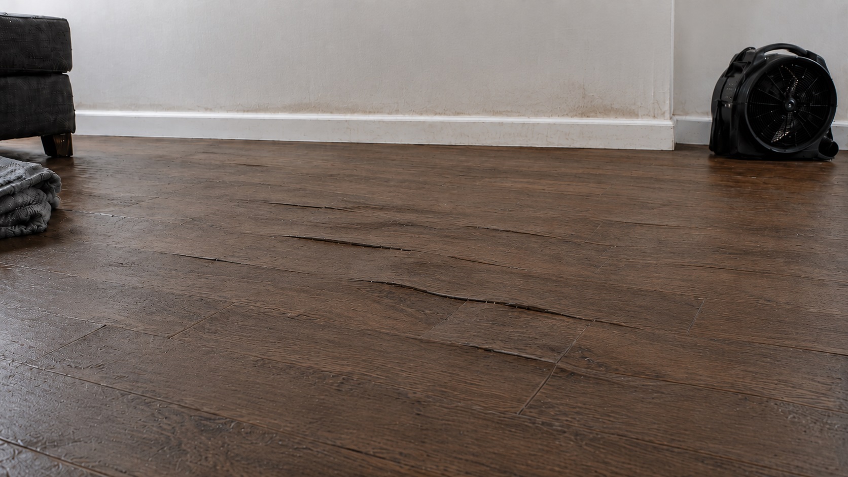 Flood-damaged laminate flooring with swollen seams, lifted planks, wall staining, a towel, and a drying fan in a partially cleared room.