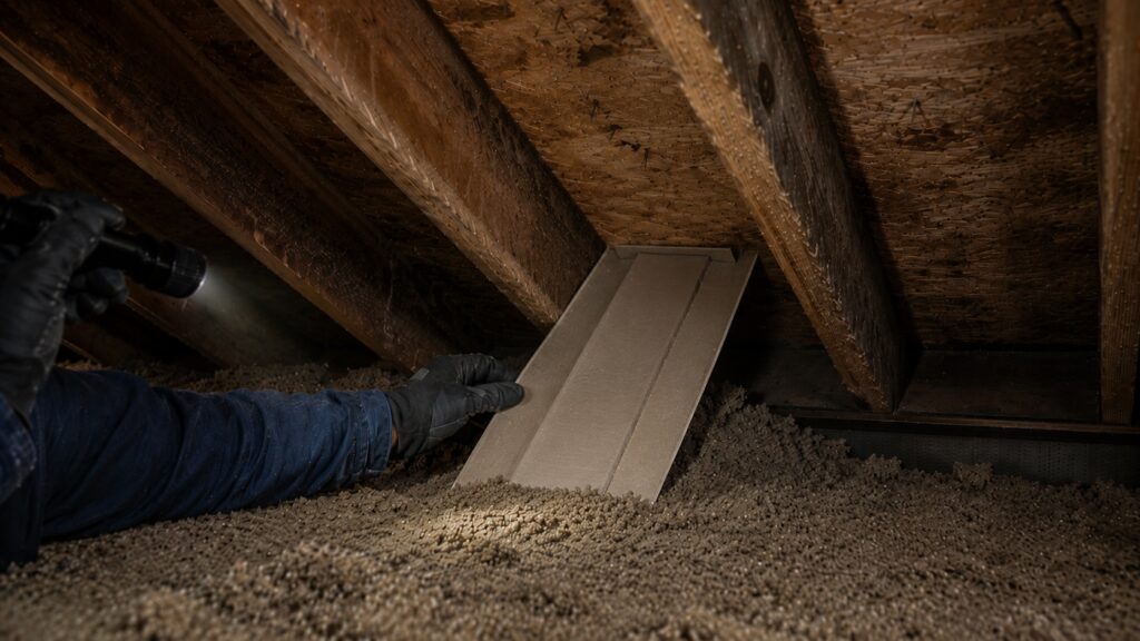 A gloved worker adjusts an attic ventilation baffle between aged rafters to keep loose insulation from blocking intake airflow.