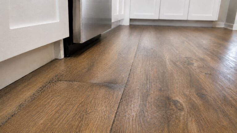 Wood-look kitchen floor near an appliance with subtle swelling, dark seam discoloration, and early mold speckling from trapped moisture after a leak.