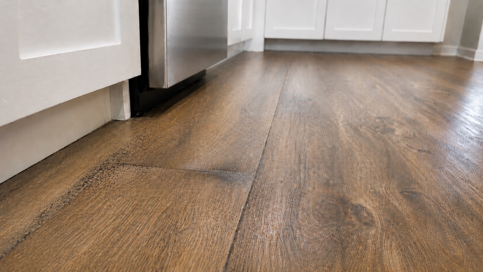 Wood-look kitchen floor near an appliance with subtle swelling, dark seam discoloration, and early mold speckling from trapped moisture after a leak.