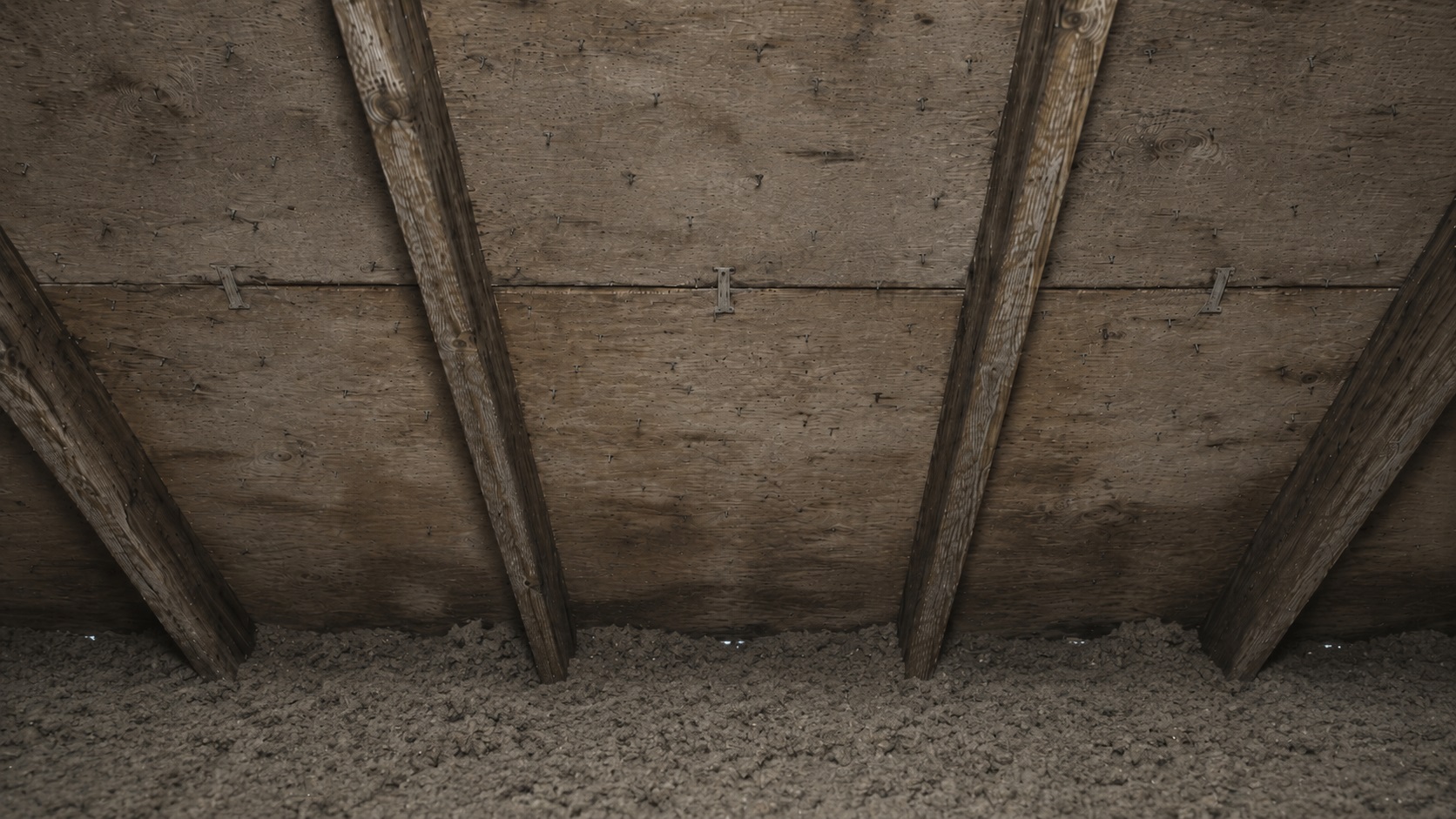 Aged attic roof decking with visible nail tips, rafters, insulation packed near the soffit, and faint moisture staining from poor ventilation.