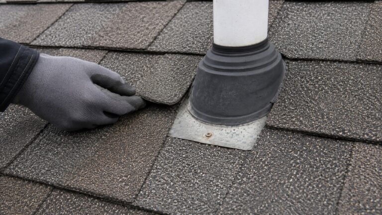 Gloved hand lifting asphalt shingles near a worn roof vent boot during a leak damage inspection.