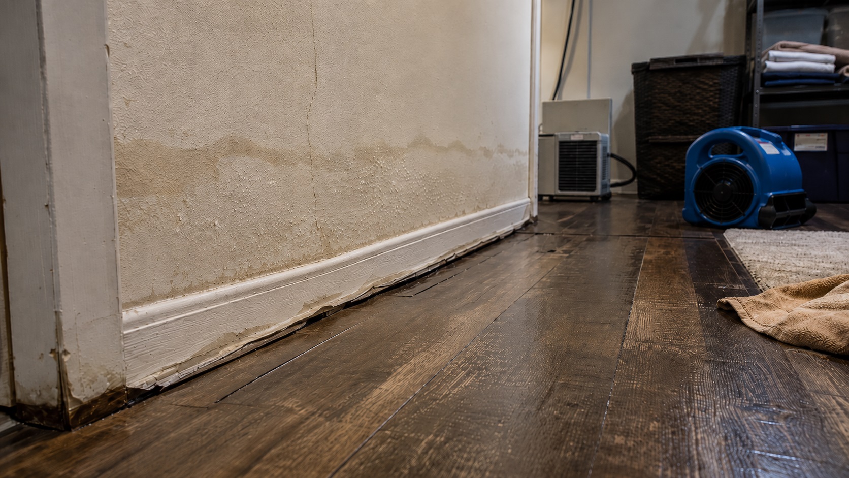 Low-angle view of a post-flood room with stained lower walls, swollen baseboards, buckled flooring, and drying equipment.