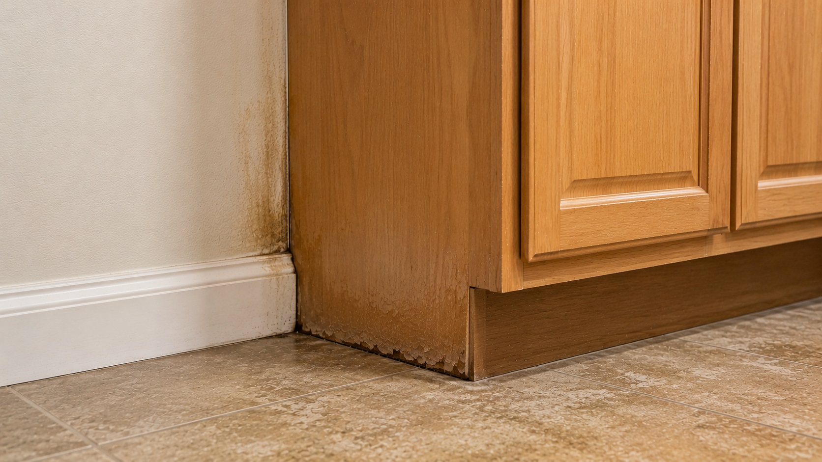 Low-angle view of a kitchen cabinet corner with subtle water staining, swollen wood, and darkened seams near the floor and wall.