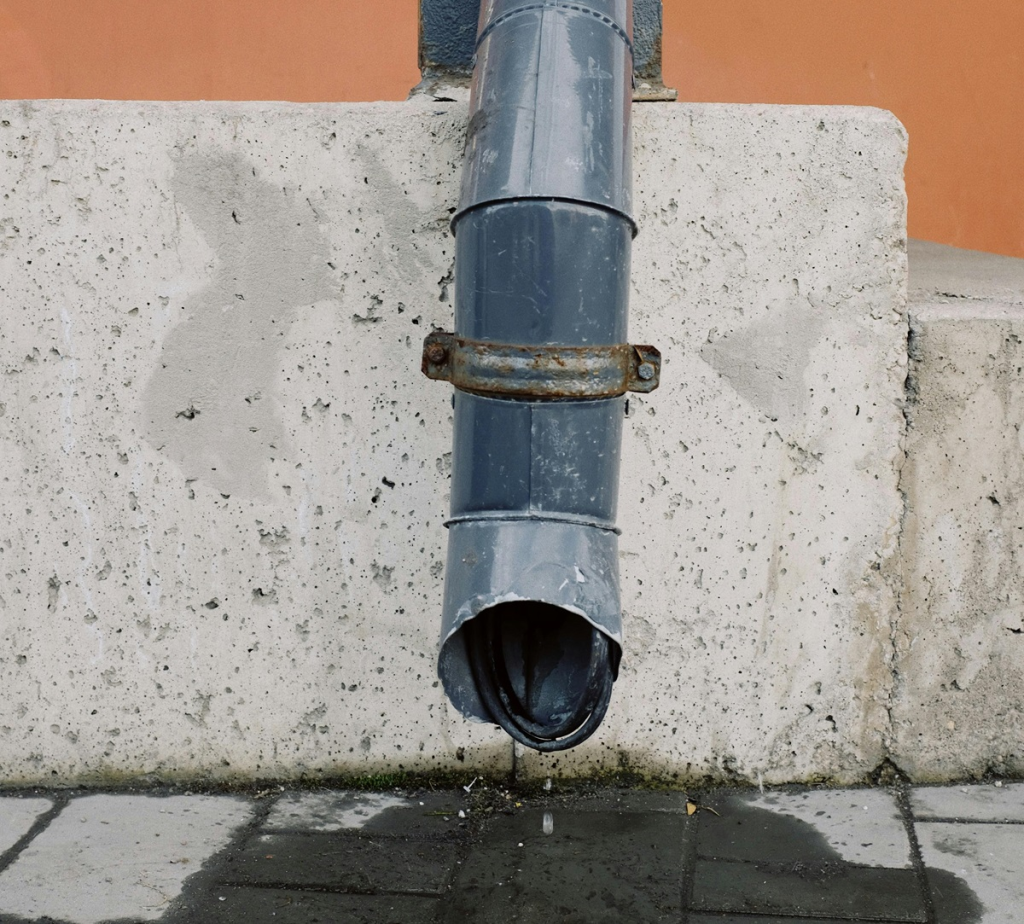 Leaking downspout pipe releasing water onto paving beside an exterior wall, showing how roof runoff can collect near a foundation.