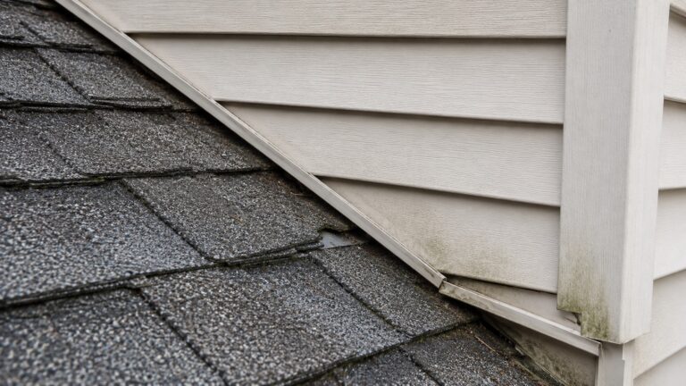 Roof-to-wall flashing problem where a broken shingle edge exposes hidden metal flashing near vinyl siding and J-channel.