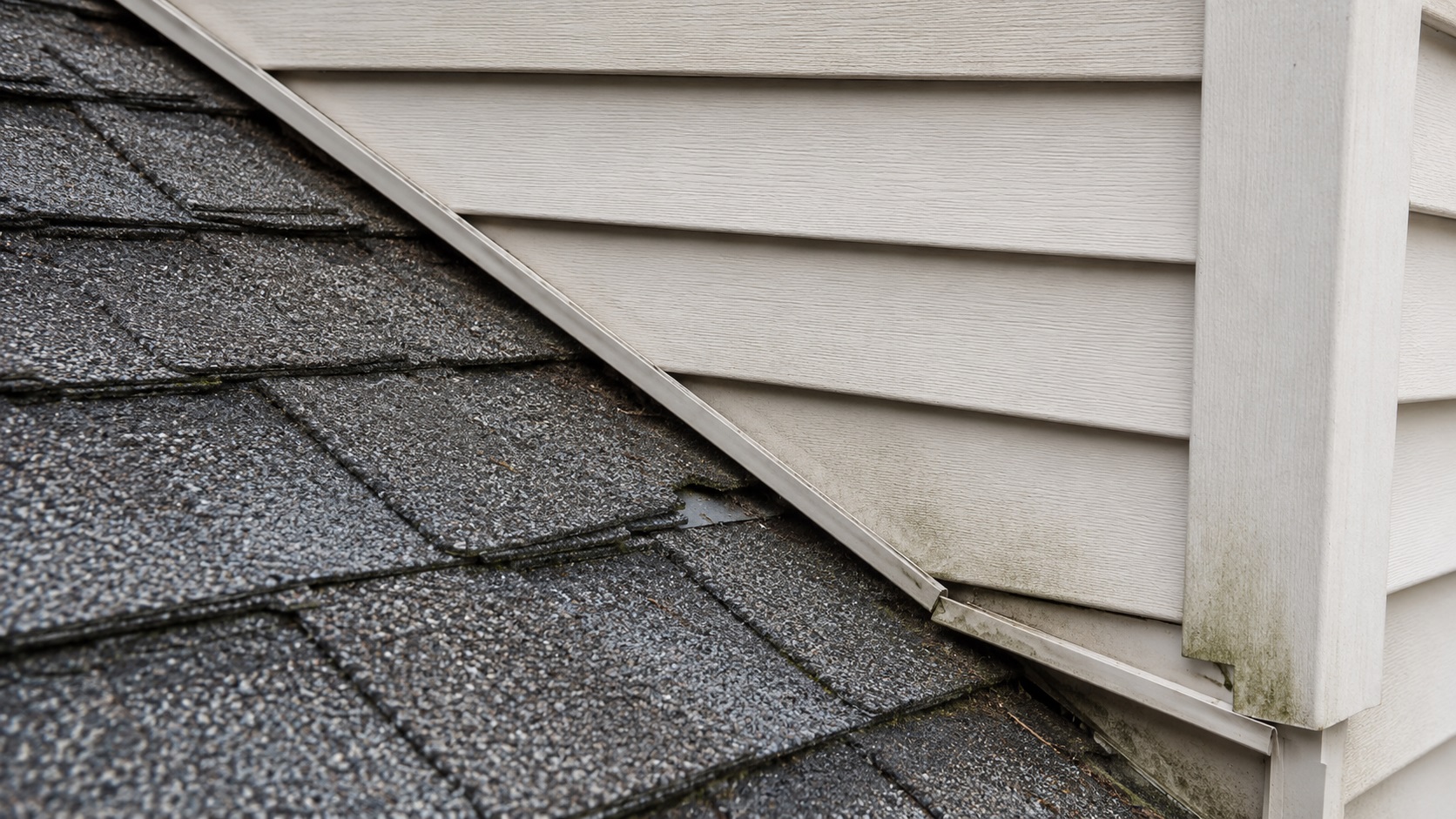 Roof-to-wall flashing problem where a broken shingle edge exposes hidden metal flashing near vinyl siding and J-channel.